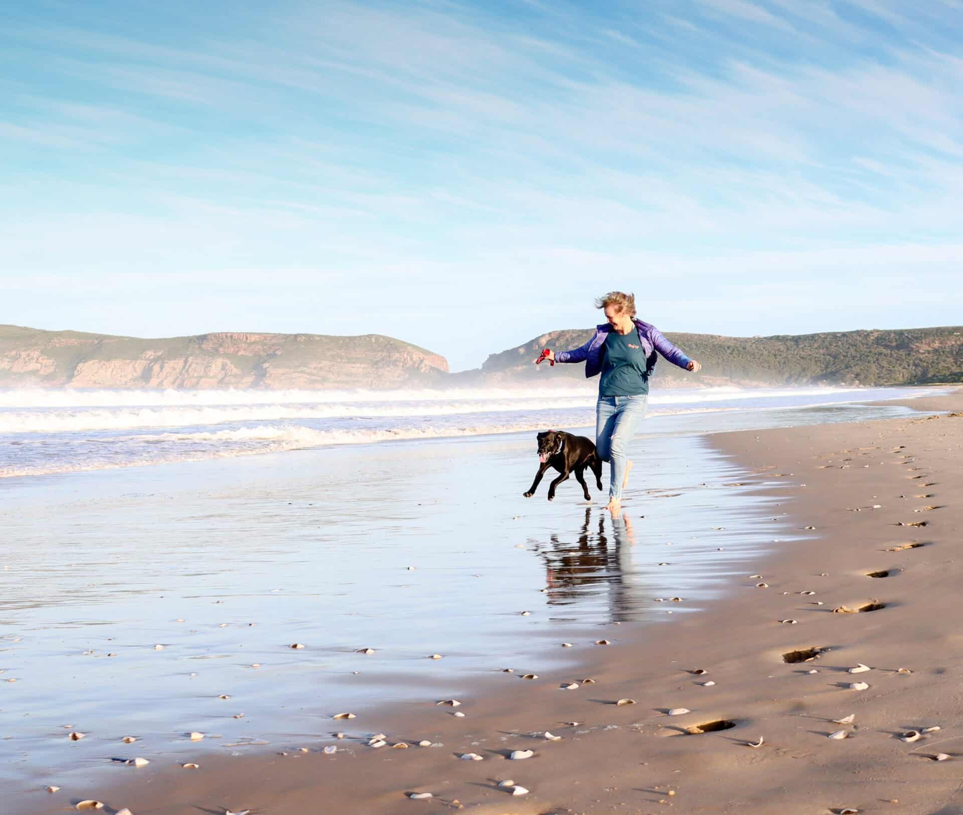 a woman running along Robberg beach with her black dog